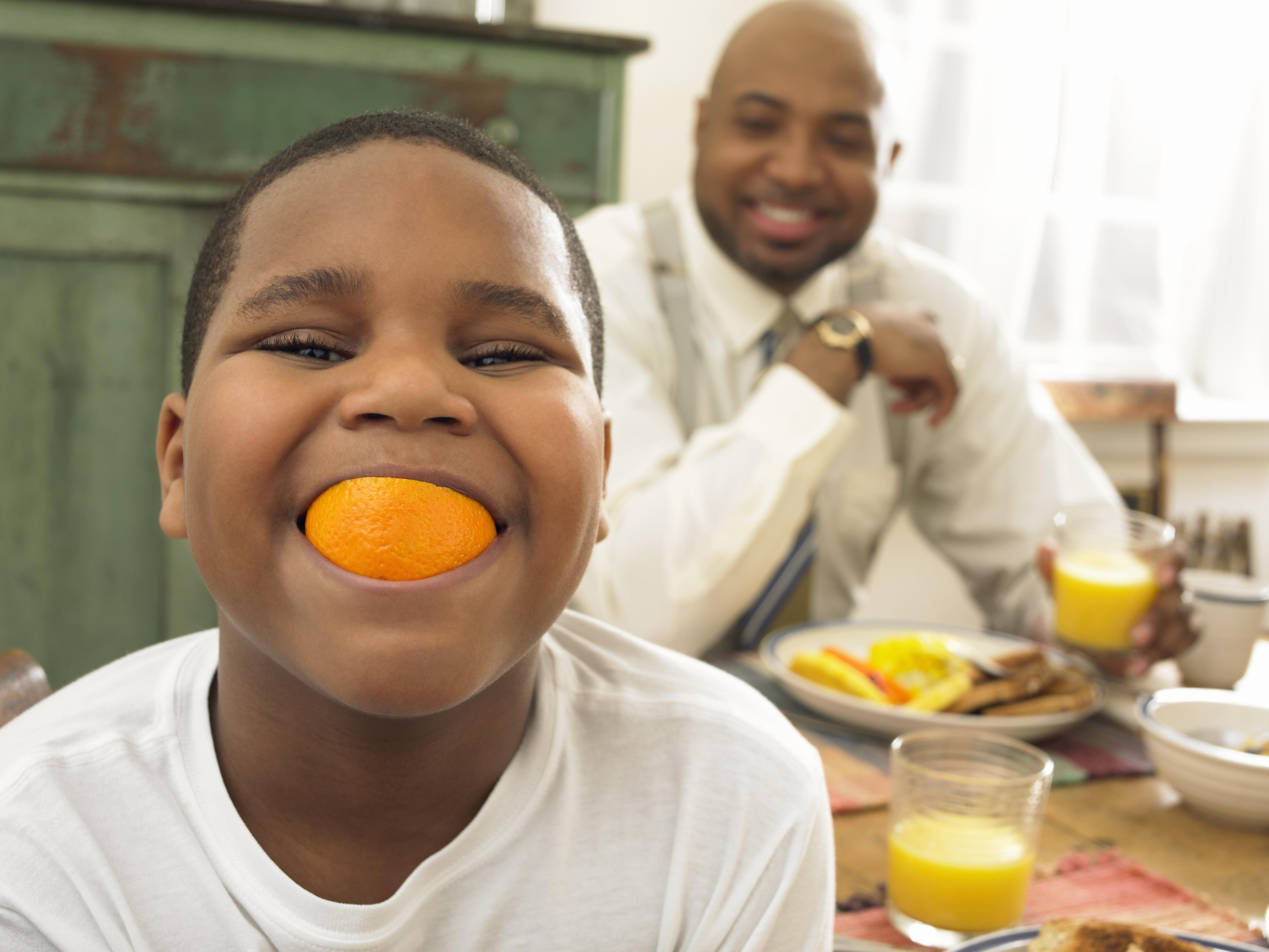 Boy With an Orange in His Mouth at Breakfast and His Father in the Background