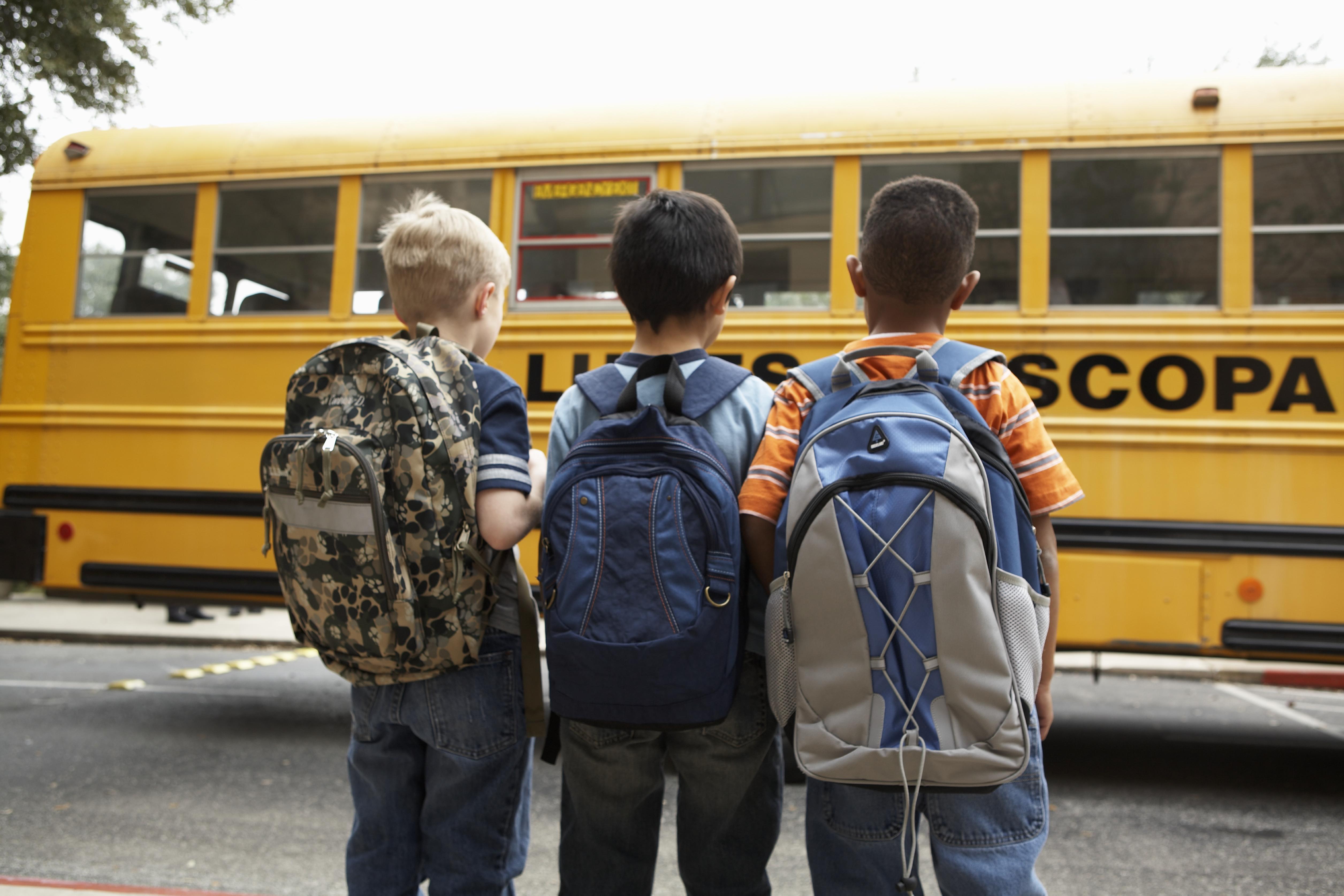 Three boys (8-9) standing with backpacks outdoors near school bus, rear view