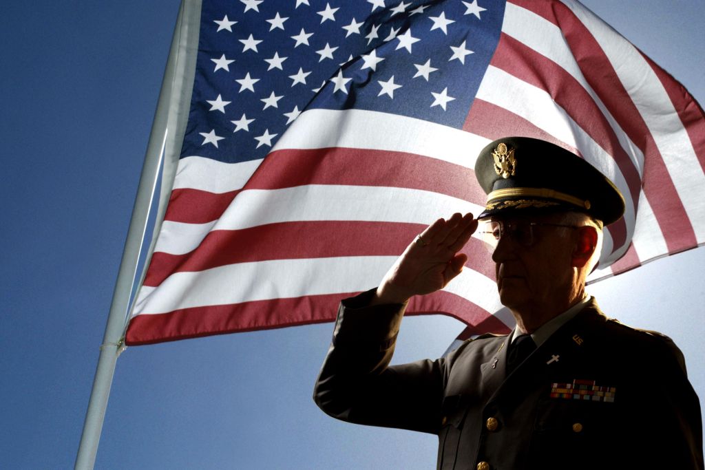 Silhouette of veteran US Army Colonel Chaplain wearing hat and saluting with an American flag flying behind him.