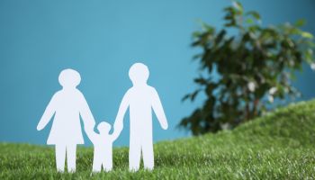 Paper cutout of parents with their child on green grass against light blue background, space for text. Family Day