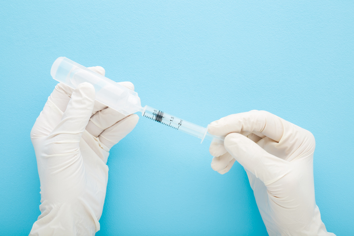 Doctor hands in white rubber protective gloves holding syringe and plastic ampoule with drug liquid. Blue table background. Vaccination and healthcare. Closeup. Point of view shot. Top down view.