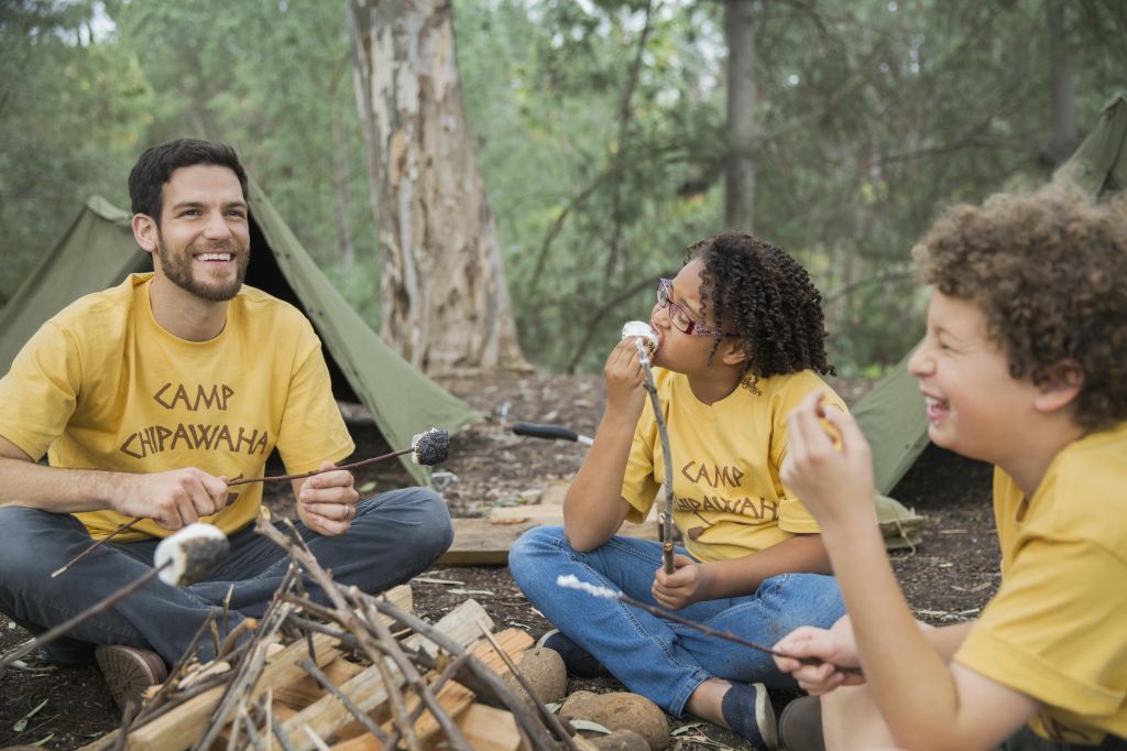Happy camp counsellor and children with marshmallows around campfire