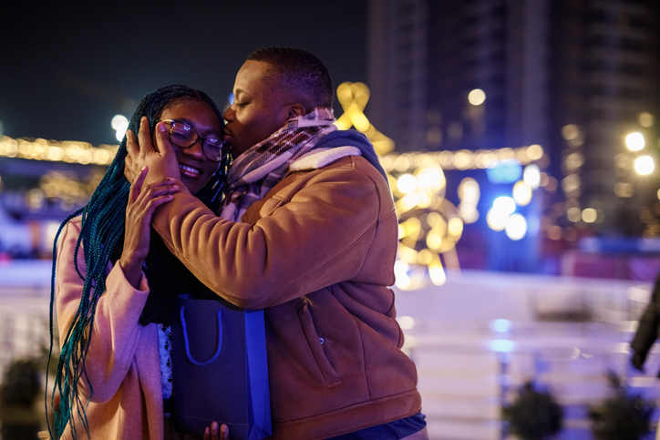 Couple shares a warm kiss and hug at night in a city setting with festive lights and a gift bag