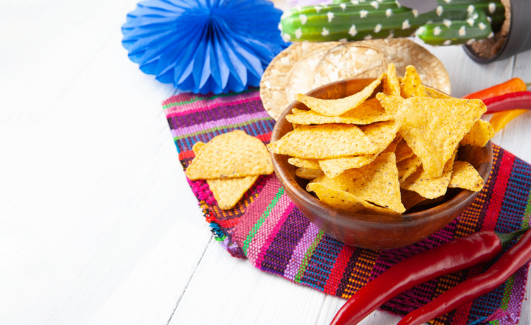 Cinco de Mayo festive composition with tortilla chips, chili peppers, cactus with sombrero and mustache and traditional Mexican textile on white wooden background