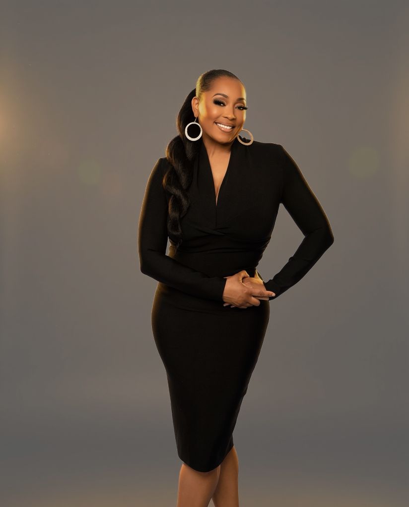 A smiling woman in a black dress with large hoop earrings stands confidently against a gray background.
