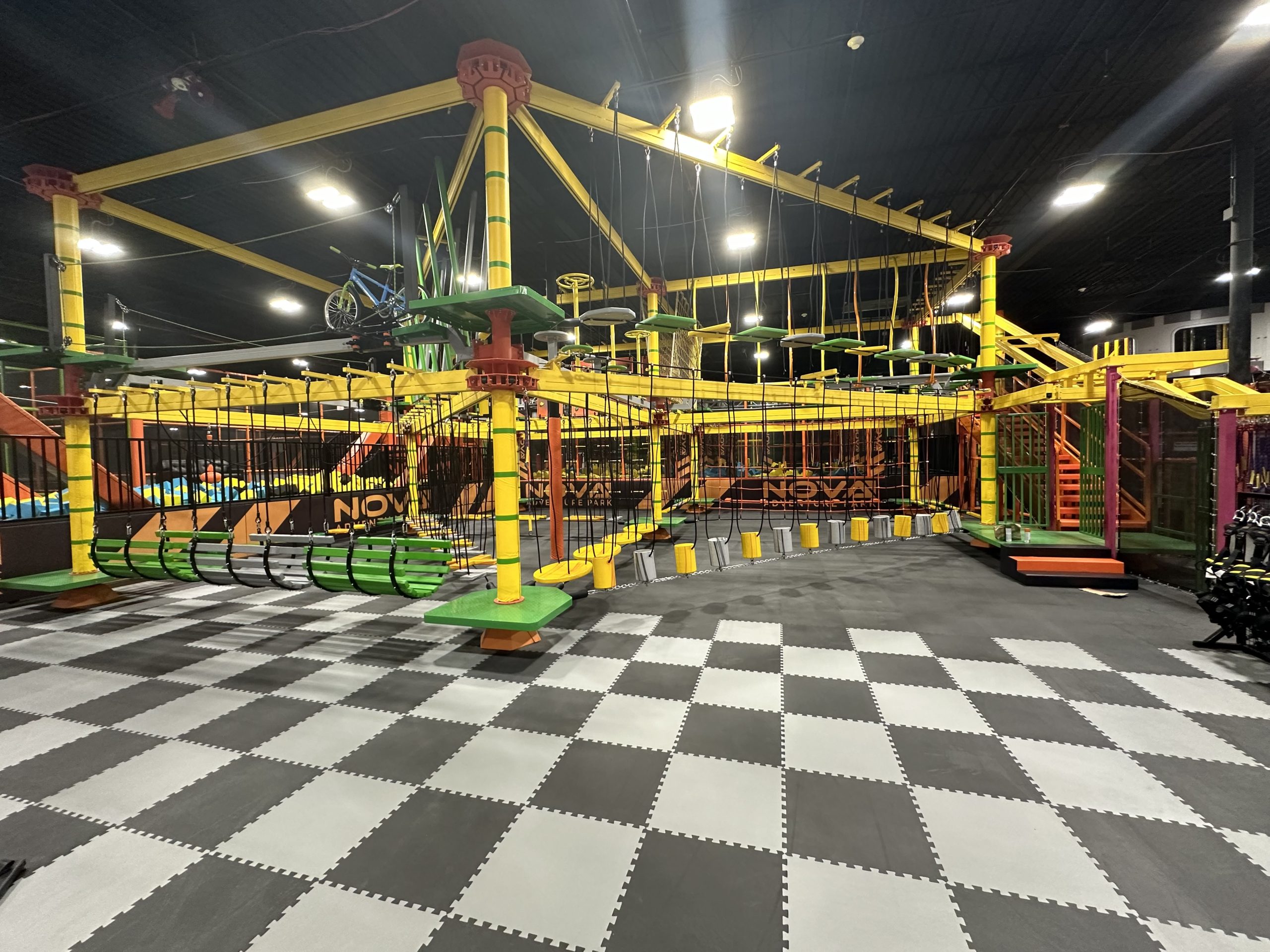 A large indoor playground with colorful climbing structures, slides, and a checkered floor. The equipment is primarily yellow and green in color.