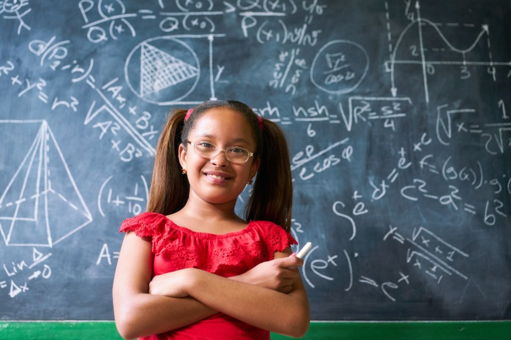 A smiling young girl in a red dress stands in front of a chalkboard covered in mathematical equations and diagrams.