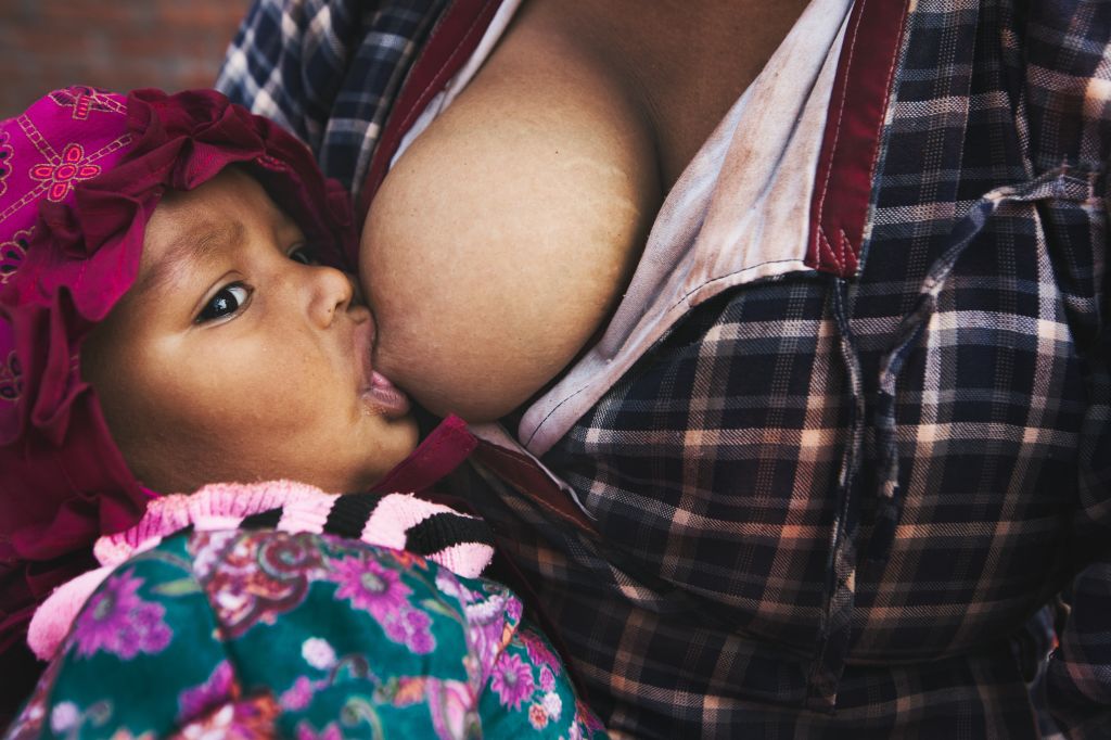 A baby nursing at their mother's breast, wearing a pink floral headband and wrapped in a plaid blanket.